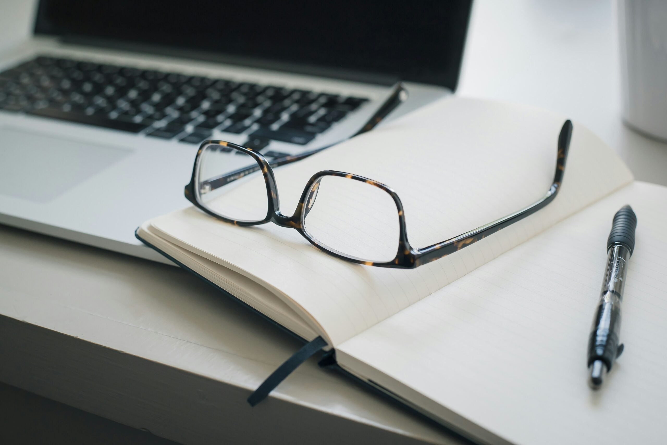 an open notebook with eyeglasses resting on top sits next to a laptop