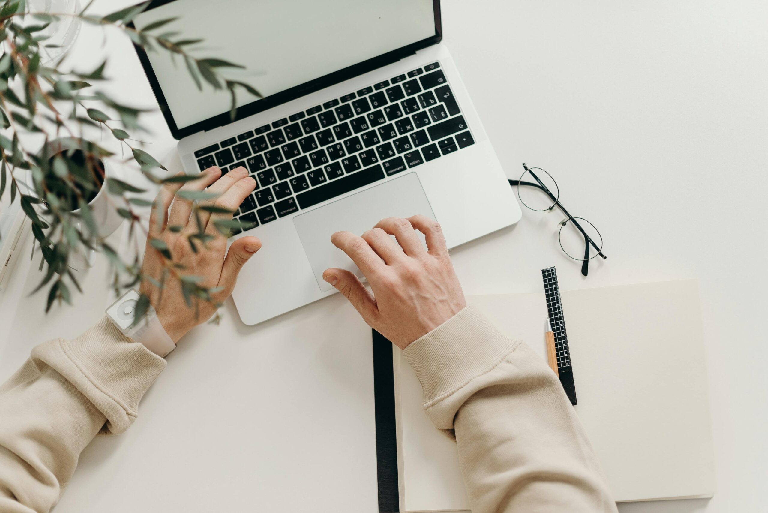 Hands typing on a laptop with a notebook, pen, and glasses on a white desk